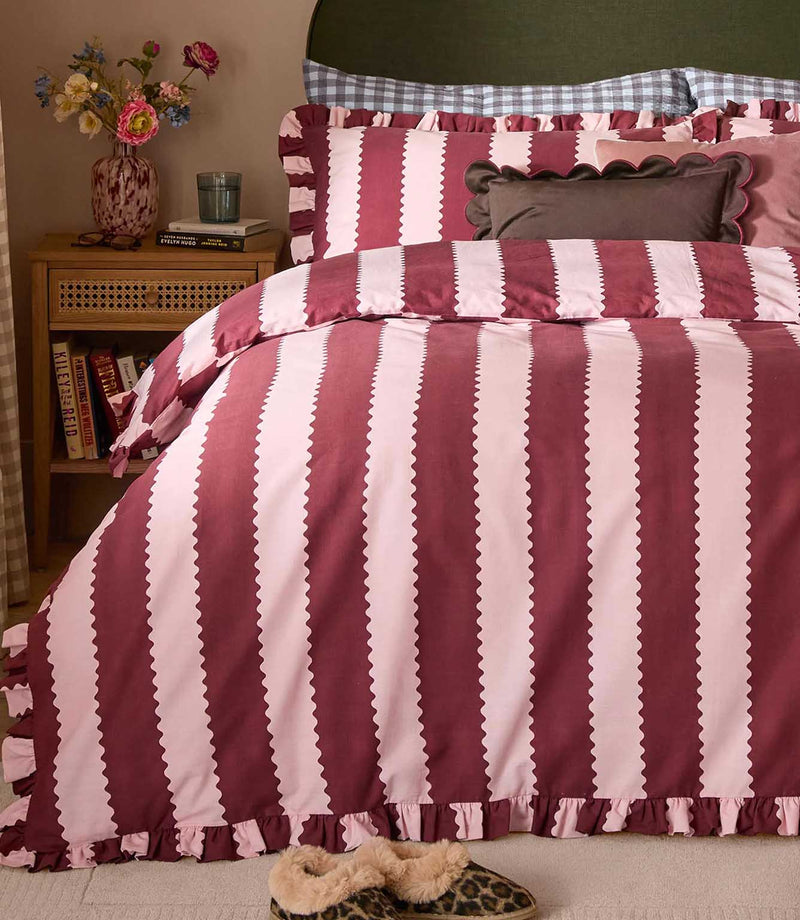 Birdie Stripe Ruffle duvet cover set in burgundy and blush pink, styled with matching striped pillowcases, ruffled edges and layered decorative cushions in a cosy bedroom setting.