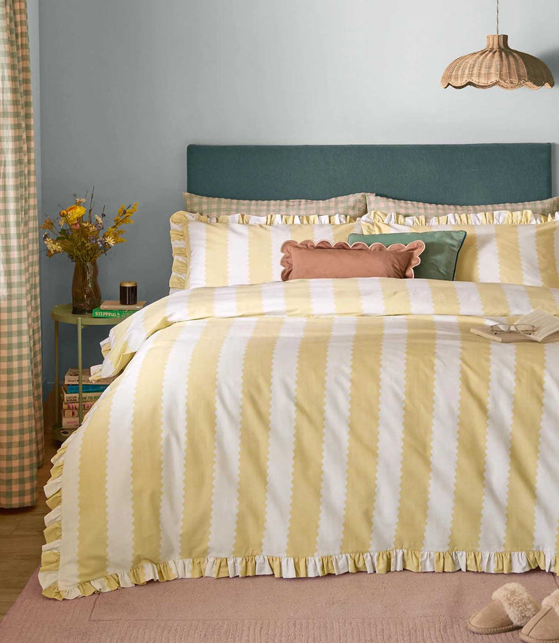 Birdie Stripe Ruffle duvet cover set in butter yellow and white, featuring wide scalloped vertical stripes and gathered ruffle edging, styled with matching pillowcases and decorative cushions.