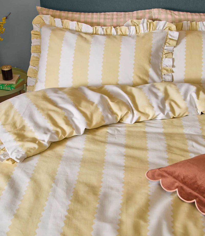 Close-up of the Birdie Stripe Ruffle bedding in butter yellow and white, showing the scalloped stripe design, soft fabric texture and gathered ruffle trim on the coordinating pillowcases.