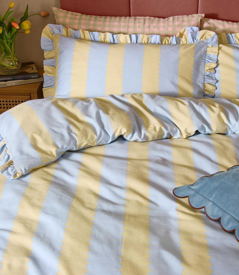 Close-up of Birdie Stripe Ruffle bedding in lemon yellow and sky blue, showing the scalloped stripe pattern, soft fabric texture and gathered two-tone ruffle trim on the pillowcases.