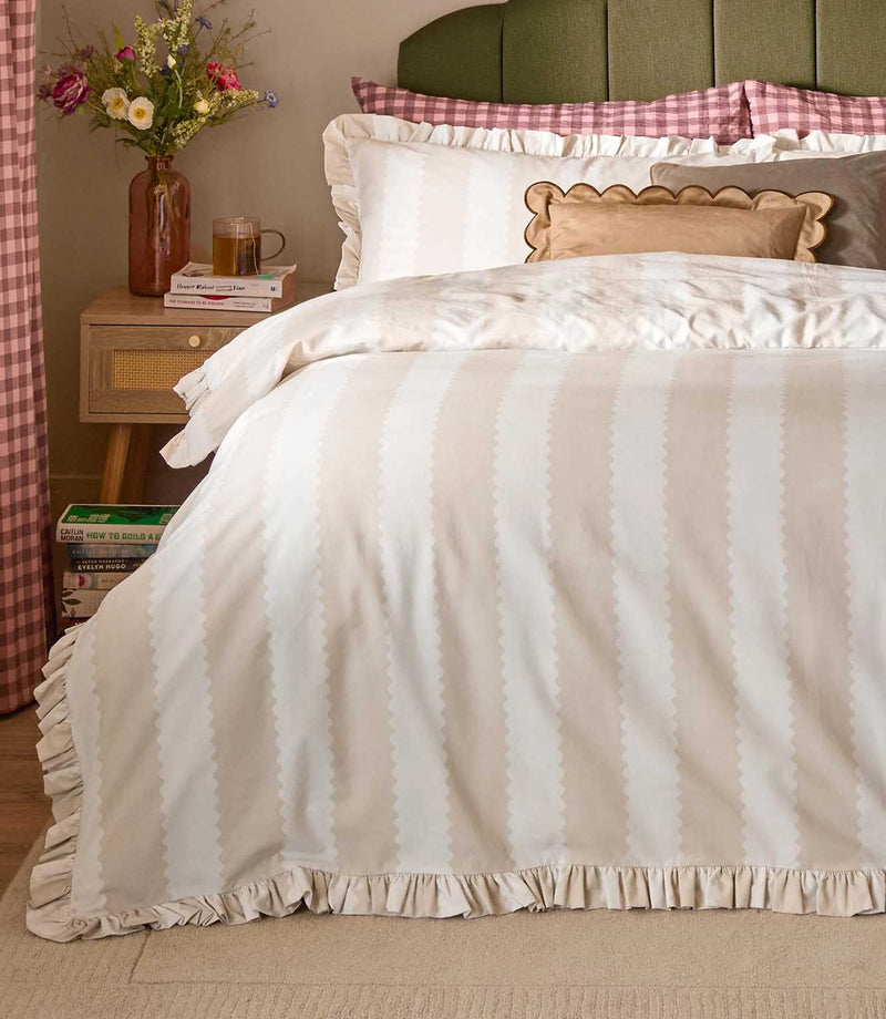 Birdie Stripe Ruffle duvet cover set in oat beige and soft white, styled on a bed with matching ruffle pillowcases and neutral decorative cushions in a warm, relaxed bedroom.