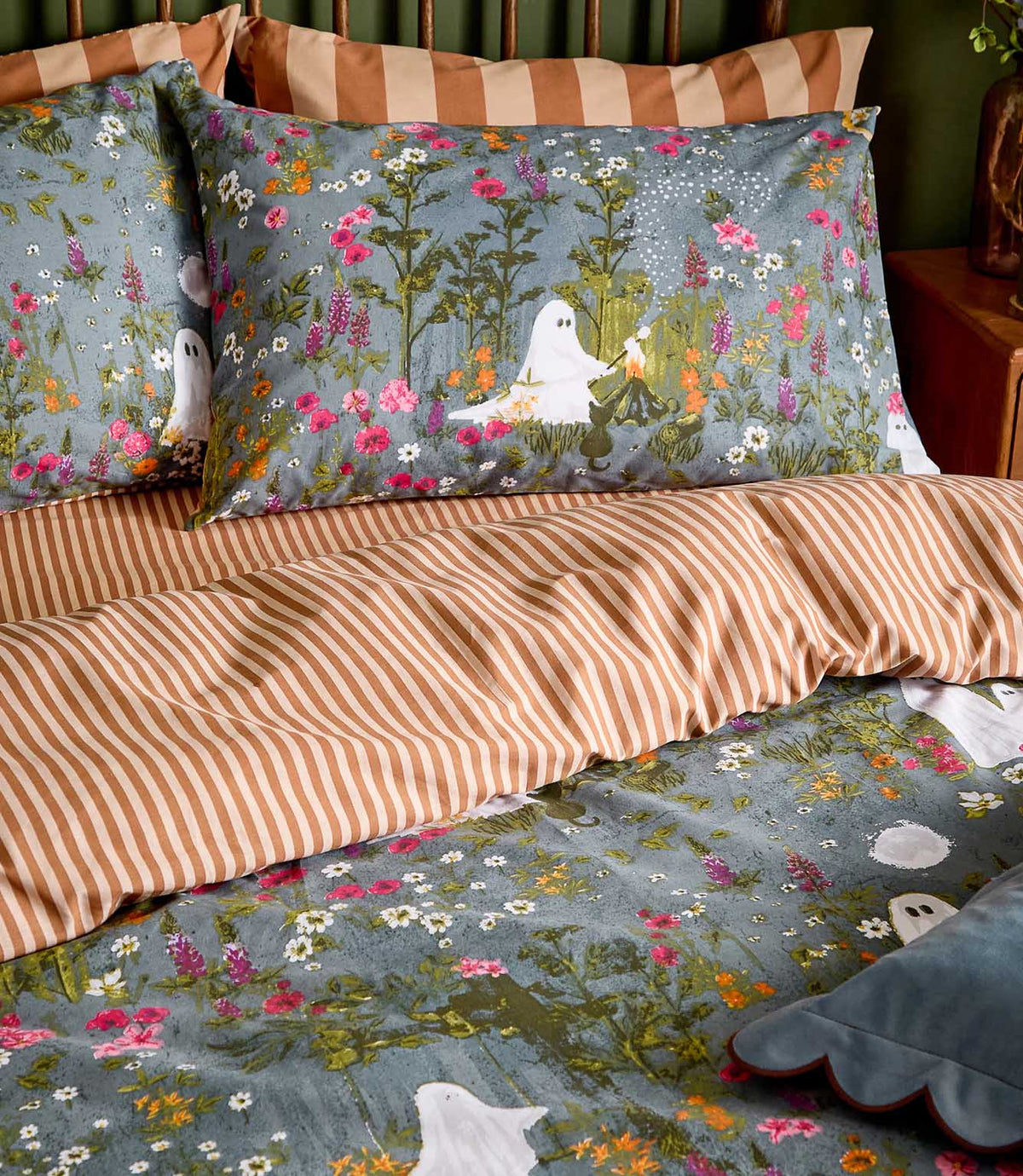 Close-up of the Bootanical Forest reversible bedding, showing white ghost and wildflower motifs on a blue-grey ground alongside the rust and cream striped reverse.