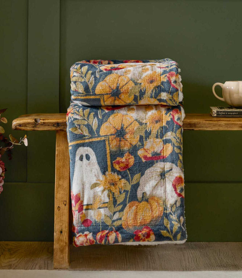 Bootanical Sherpa fleece throw folded over a rustic wooden bench, showing a dark teal print with white ghosts, orange pumpkins and autumn flowers against a green panelled wall.