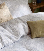 Close-up of Braeflower muslin bedding by Voyage Maison, showing the soft grey botanical pattern, relaxed crinkled texture and layered cushions.