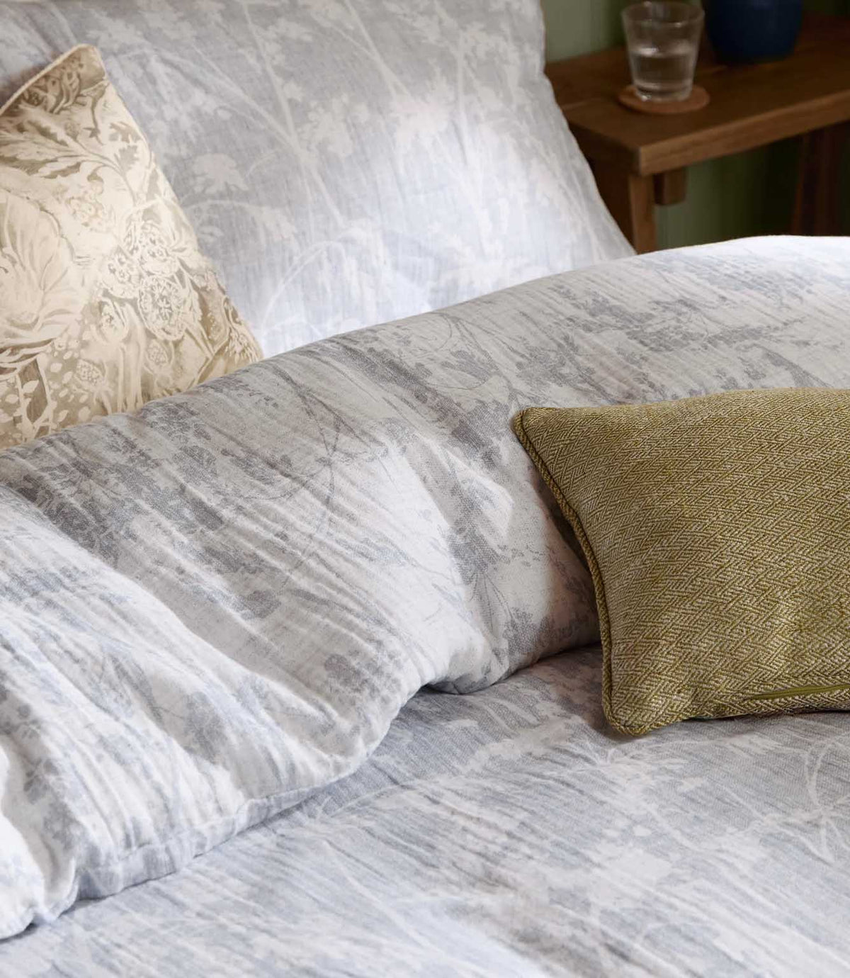 Close-up of Braeflower muslin bedding by Voyage Maison, showing the soft grey botanical pattern, relaxed crinkled texture and layered cushions.