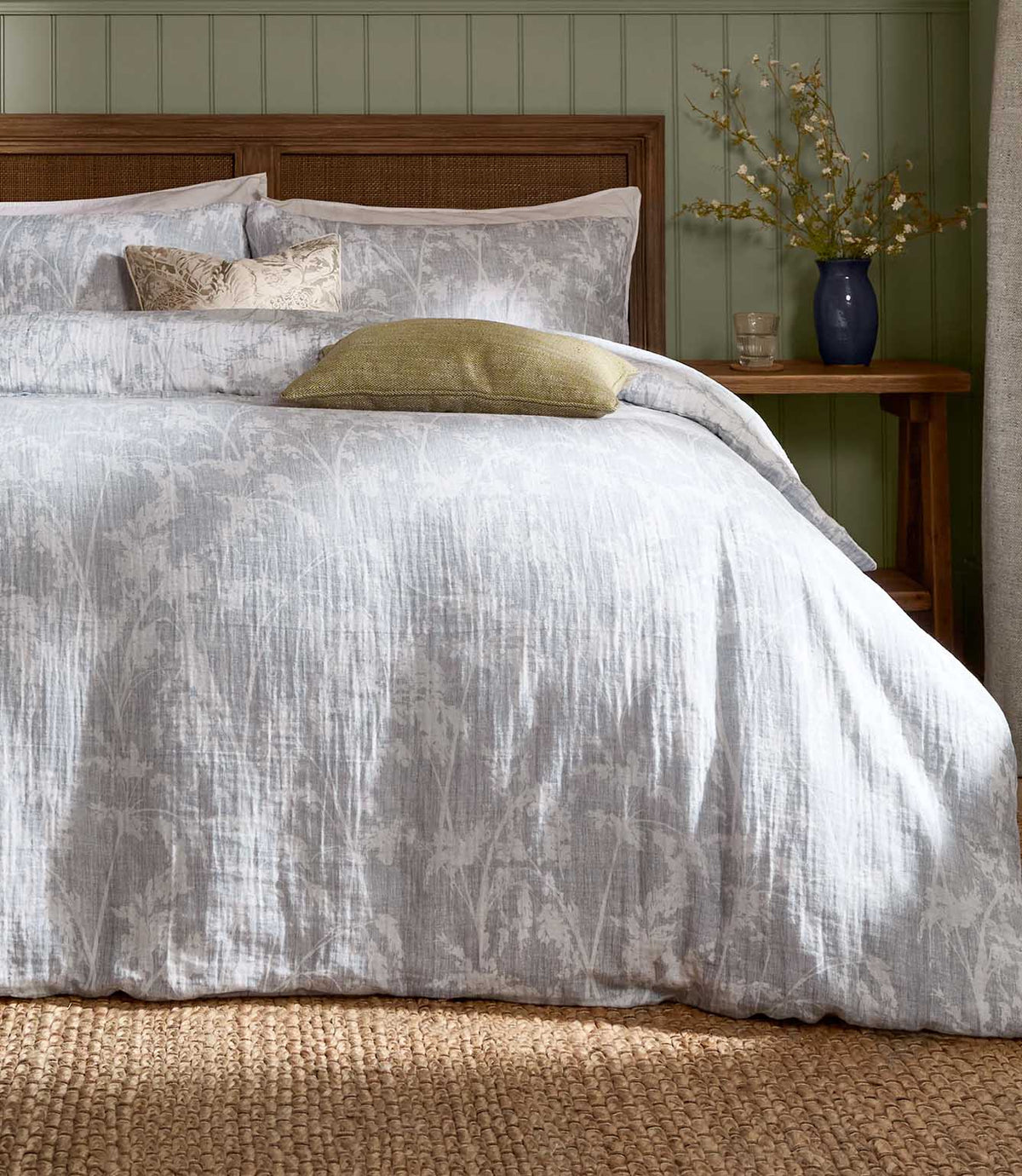 Voyage Maison Braeflower Reversible Muslin Duvet Cover Set in soft grey, styled with green cushions against a wooden headboard and sage panelling.