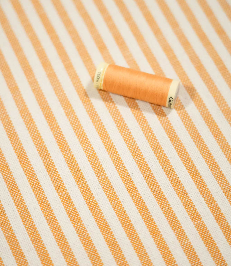 Broadway Stripe Sunflower fabric displayed with a coordinating orange thread spool, showing the warm sunflower orange and off-white vertical stripe design and the visible woven texture of the fabric.
