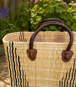 A woven straw tote basket with dark brown leather handles and vertical black stripe detailing on the front, photographed outdoors in front of a garden background.