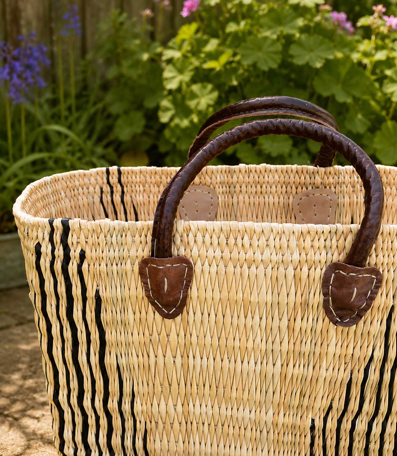 A woven straw tote basket with dark brown leather handles and vertical black stripe detailing on the front, photographed outdoors in front of a garden background.