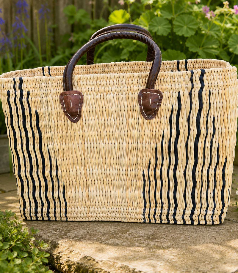 A woven straw tote bag with black vertical stripes and dark brown leather handles, resting on a stone surface with a garden background.
