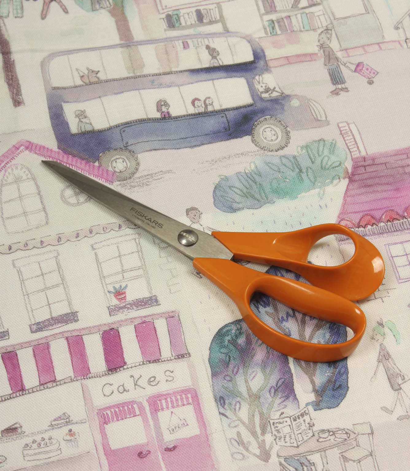 Cakes & More cotton fabric displayed with scissors, highlighting the playful townscape print in purple and pink shades.