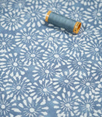Chamberry Bluebell fabric photographed with a coordinating blue sewing thread spool for scale, showing the small white floral motifs, muted blue background and subtly textured woven surface.