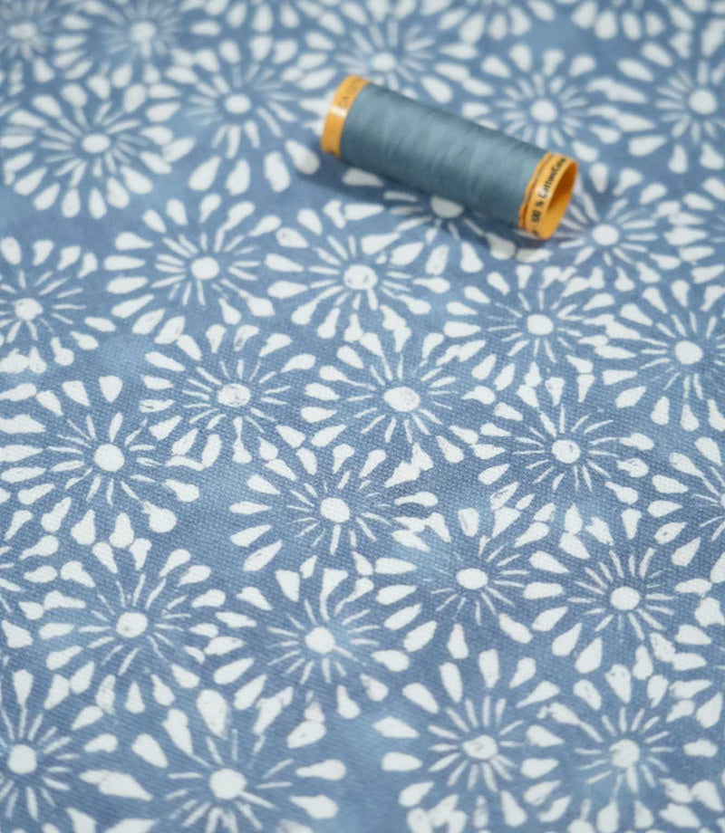 Chamberry Bluebell fabric photographed with a coordinating blue sewing thread spool for scale, showing the small white floral motifs, muted blue background and subtly textured woven surface.