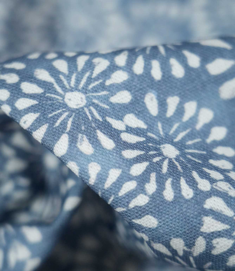 Macro photograph of Chamberry Bluebell fabric showing the fine woven texture and detailed white floral motifs, with rounded petals and delicate radiating lines printed onto a soft blue background.