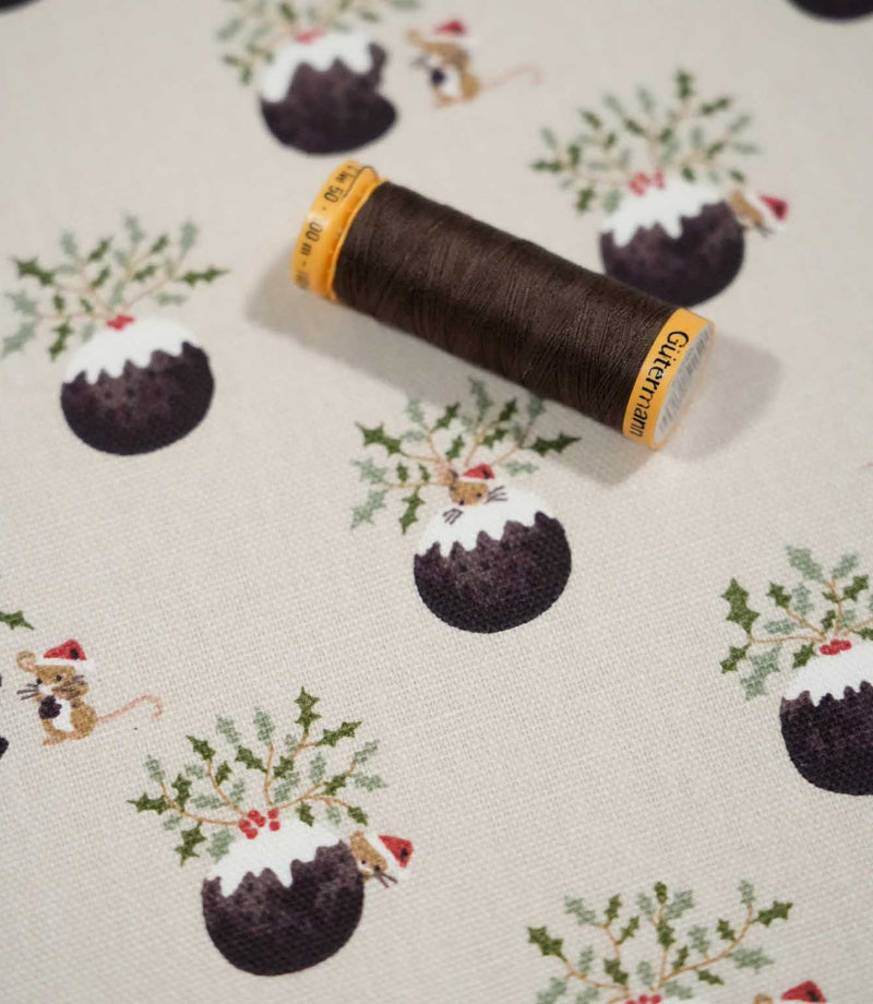 Christmas Pudding Neutral festive fabric with a coordinating brown thread spool, showing Christmas pudding, holly and mouse motifs on a neutral woven ground.