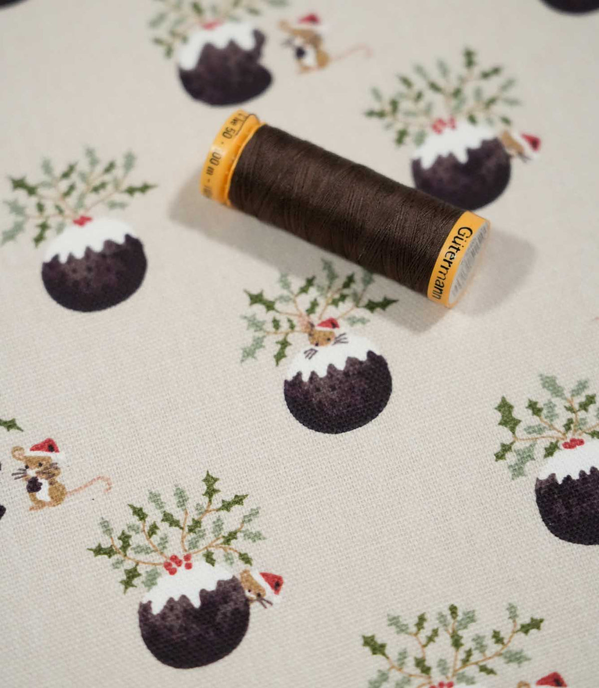 Christmas Pudding Neutral festive fabric with a coordinating brown thread spool, showing Christmas pudding, holly and mouse motifs on a neutral woven ground.