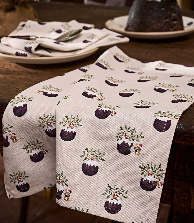 A close-up festive table runner with fabric featuring Christmas pudding illustrations with holly, mice and mistletoe on a light beige textured background.