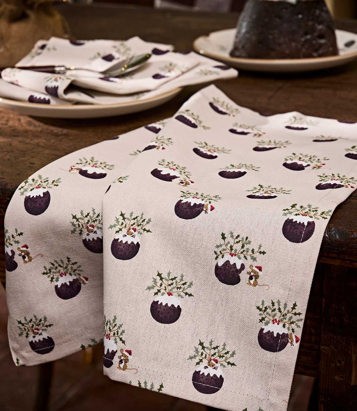 A close-up festive table runner with fabric featuring Christmas pudding illustrations with holly, mice and mistletoe on a light beige textured background.