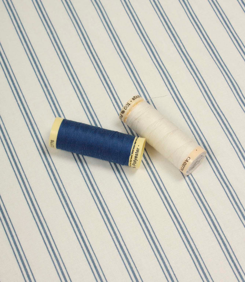 Classic striped fabric shown with navy blue and ivory thread spools against a delicate blue ticking stripe pattern.