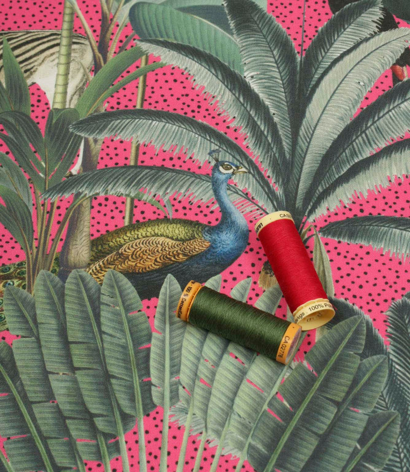 Tropical animal upholstery fabric with lush green botanical leaves, a peacock motif and vibrant pink background with coordinating thread spools.