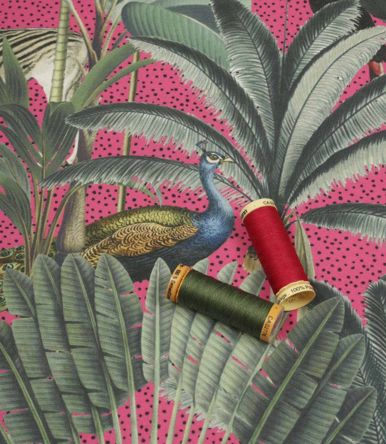 Tropical animal upholstery fabric with lush green botanical leaves, a peacock motif and vibrant pink background with coordinating thread spools.