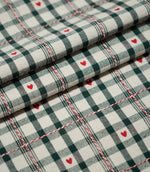 Layered folds of Cotswold Heart Tartan fabric, featuring an ivory and dark green check with small red hearts and decorative red and white accents.