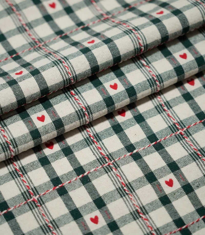 Layered folds of Cotswold Heart Tartan fabric, featuring an ivory and dark green check with small red hearts and decorative red and white accents.
