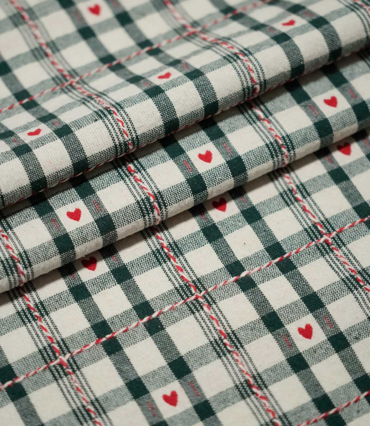 Layered folds of Cotswold Heart Tartan fabric, featuring an ivory and dark green check with small red hearts and decorative red and white accents.