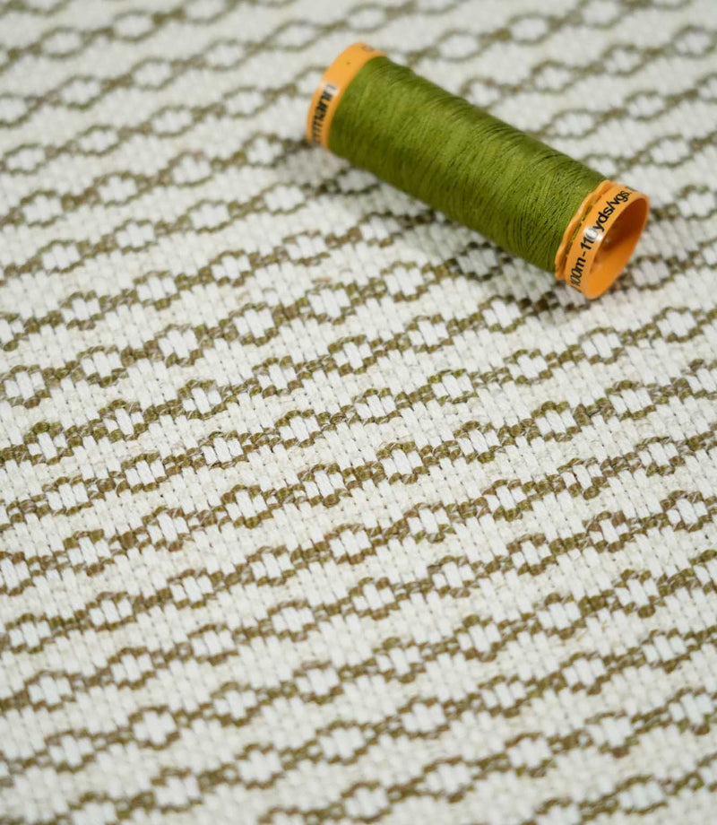 Elkstone Matcha fabric photographed with an olive green thread spool for scale, highlighting its textured cream weave and repeating olive green geometric pattern.