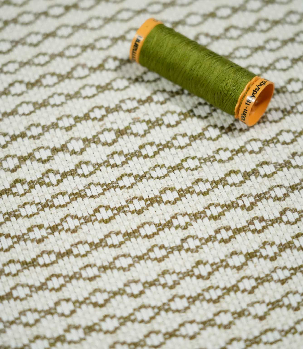 Elkstone Matcha fabric photographed with an olive green thread spool for scale, highlighting its textured cream weave and repeating olive green geometric pattern.
