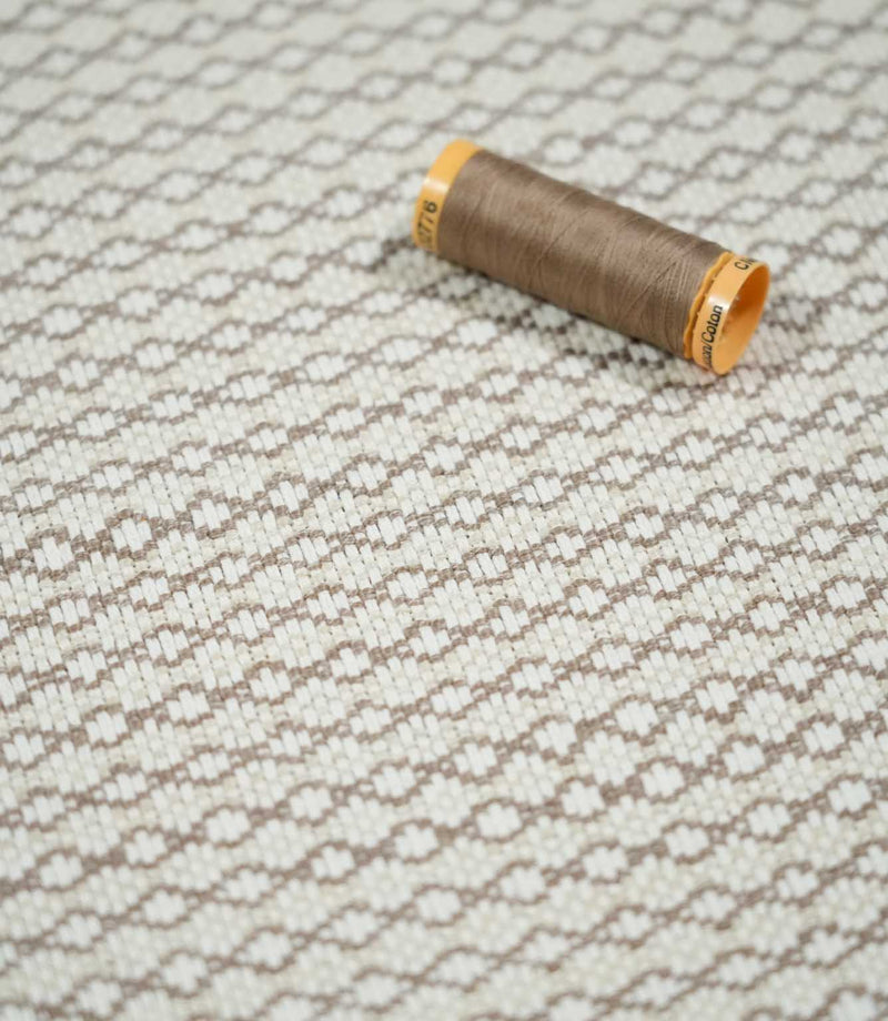 Elkstone Taupe fabric photographed with a matching taupe thread spool for scale, highlighting its textured cream weave and repeating geometric pattern.