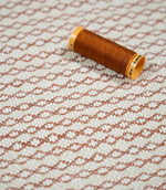 Elkstone Terracotta fabric photographed with a rust-orange thread spool for scale, highlighting its textured cream weave and repeating terracotta geometric pattern.