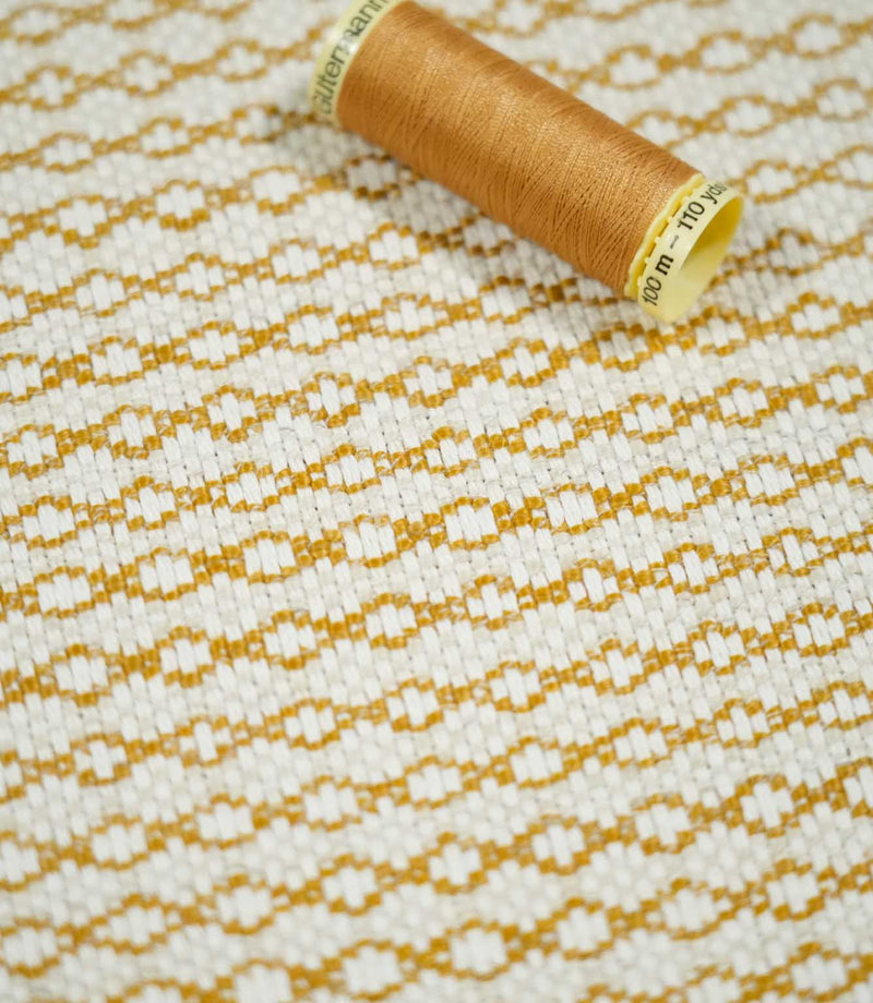 Elkstone Turmeric fabric photographed with a matching golden yellow thread spool for scale, highlighting its textured cream weave and repeating geometric pattern.