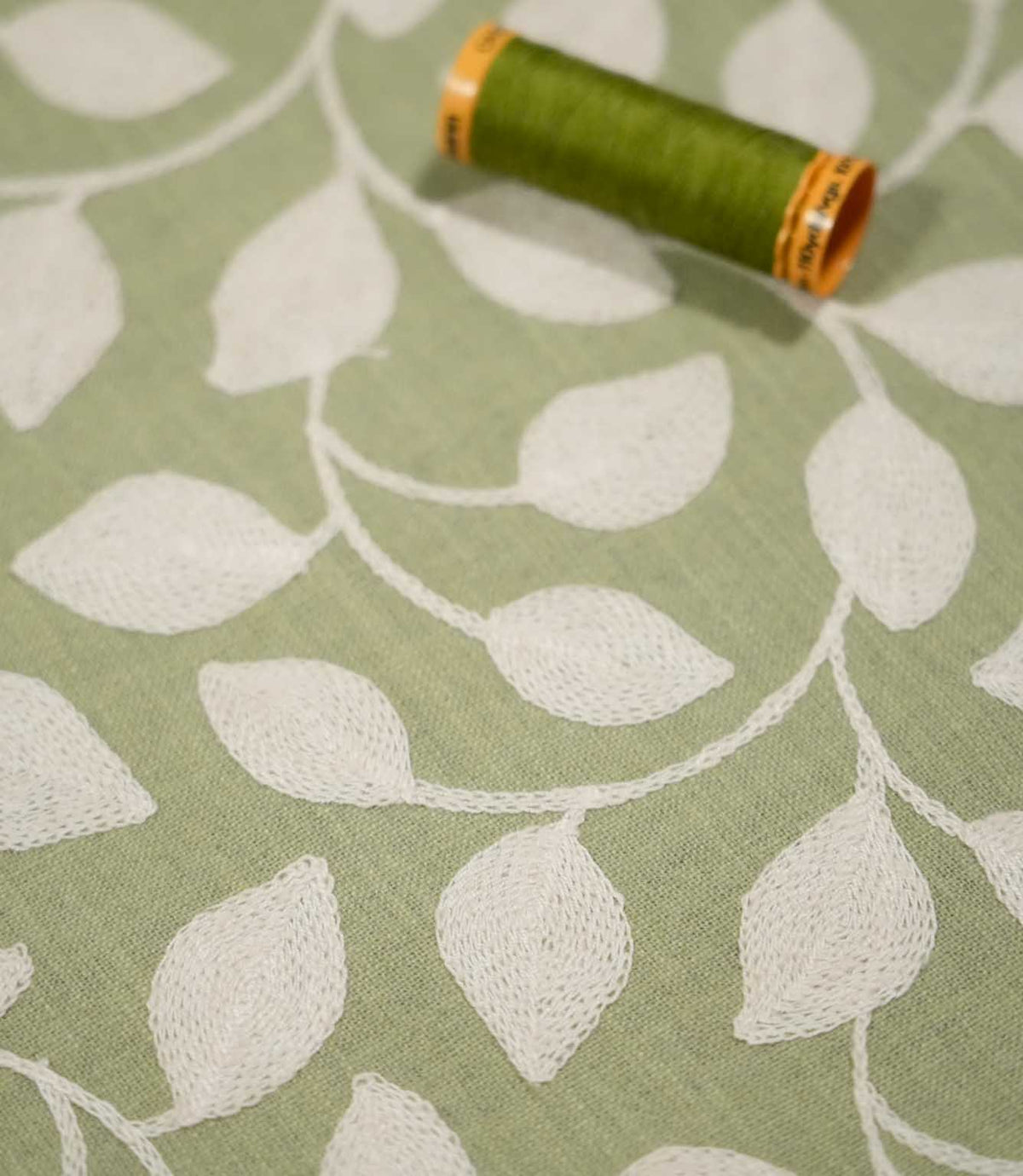 Ellerton Apple embroidered foliage fabric shown alongside a coordinating green thread spool, with textured cream leaves and winding stems across an apple green background, providing a close view of the fabric weave, embroidery and botanical detailing.