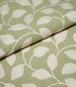 Close-up of folded Ellerton Apple fabric displaying the cream embroidered foliage pattern across an apple green woven background, with raised leaves and trailing stems continuing naturally over the fold to show the fabric's texture and structure.