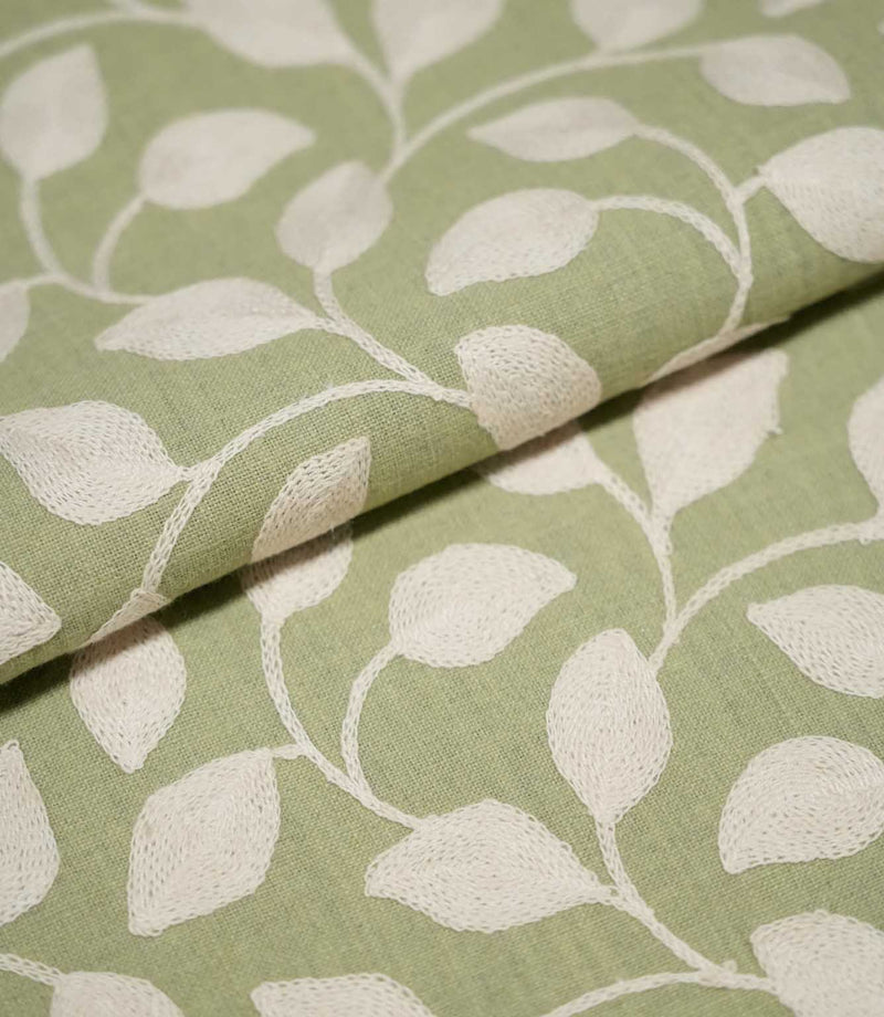 Close-up of folded Ellerton Apple fabric displaying the cream embroidered foliage pattern across an apple green woven background, with raised leaves and trailing stems continuing naturally over the fold to show the fabric's texture and structure.