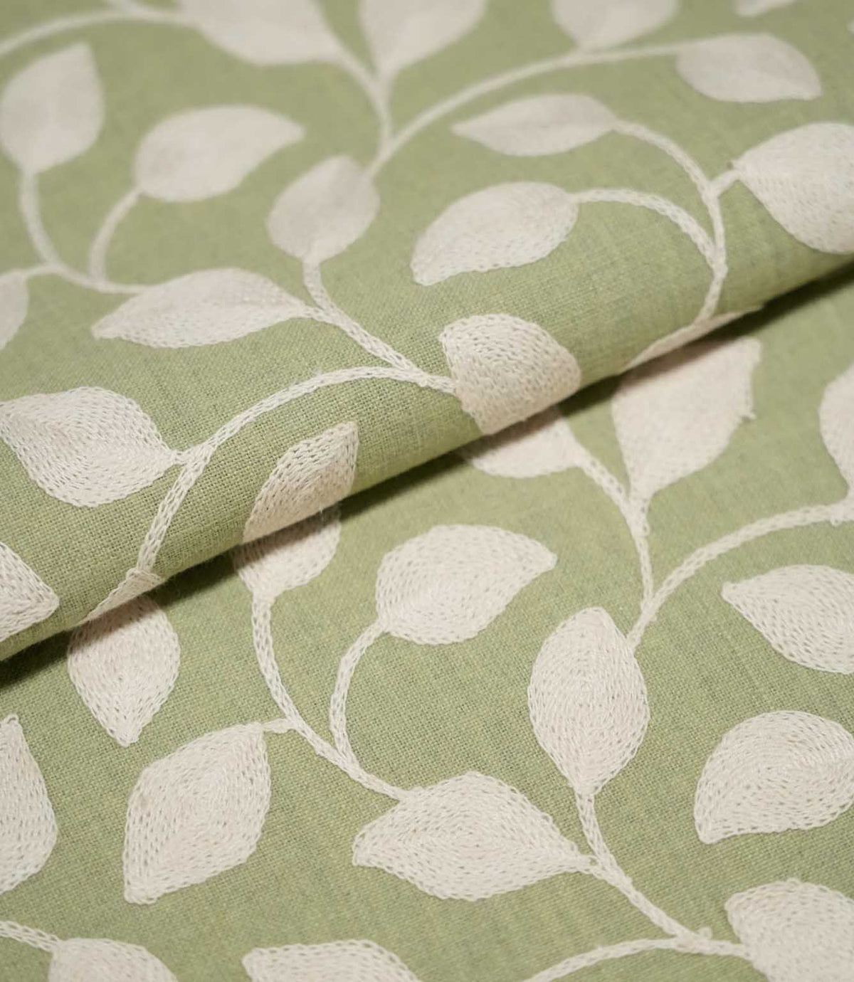 Close-up of folded Ellerton Apple fabric displaying the cream embroidered foliage pattern across an apple green woven background, with raised leaves and trailing stems continuing naturally over the fold to show the fabric's texture and structure.