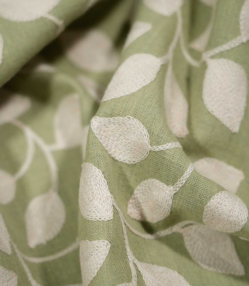Detailed close-up of Ellerton Apple fabric gathered into soft sculptural folds, focusing on the raised cream embroidered leaves, fine stitched stems and textured apple green woven background, clearly showing the depth and tactile character of this contemporary foliage fabric.
