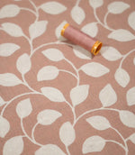 Ellerton Blush embroidered foliage fabric displayed flat with a coordinating blush pink thread spool, showing cream stitched leaves and sweeping stems across the softly textured pink background, with shallow focus emphasising the embroidery and fabric surface.