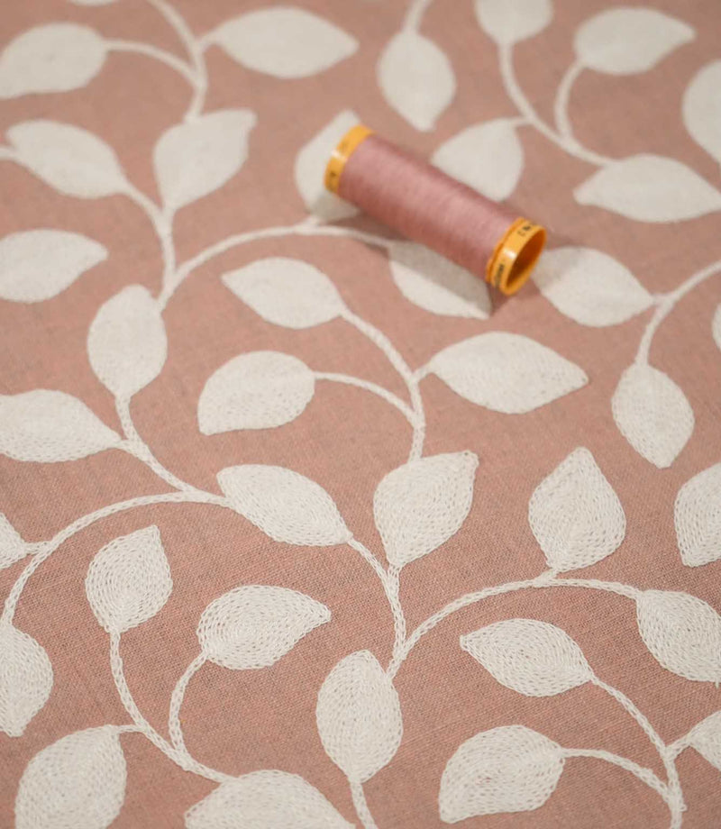 Ellerton Blush embroidered foliage fabric displayed flat with a coordinating blush pink thread spool, showing cream stitched leaves and sweeping stems across the softly textured pink background, with shallow focus emphasising the embroidery and fabric surface.