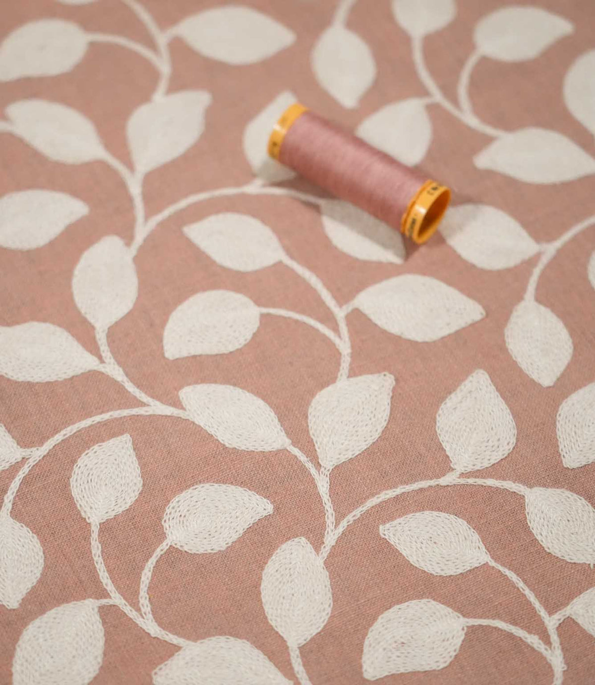 Ellerton Blush embroidered foliage fabric displayed flat with a coordinating blush pink thread spool, showing cream stitched leaves and sweeping stems across the softly textured pink background, with shallow focus emphasising the embroidery and fabric surface.