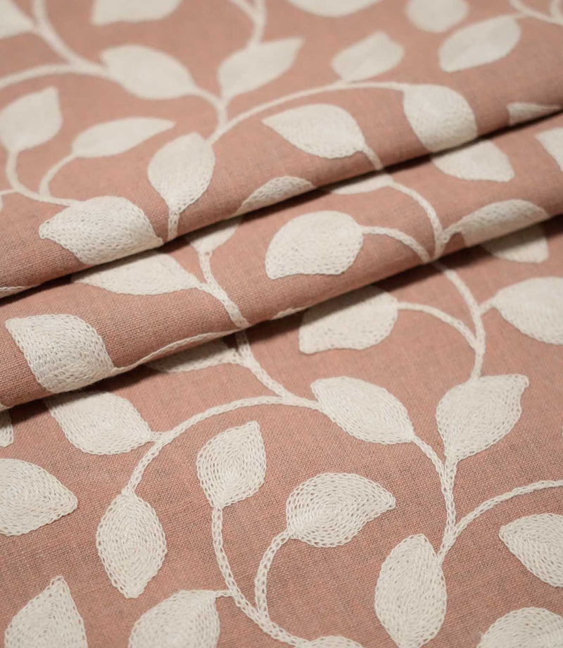Ellerton Blush embroidered fabric arranged in overlapping folds, featuring raised cream botanical leaves connected by flowing curved stems on a blush pink woven background, highlighting the repeating foliage pattern, detailed stitching and natural movement of the fabric.