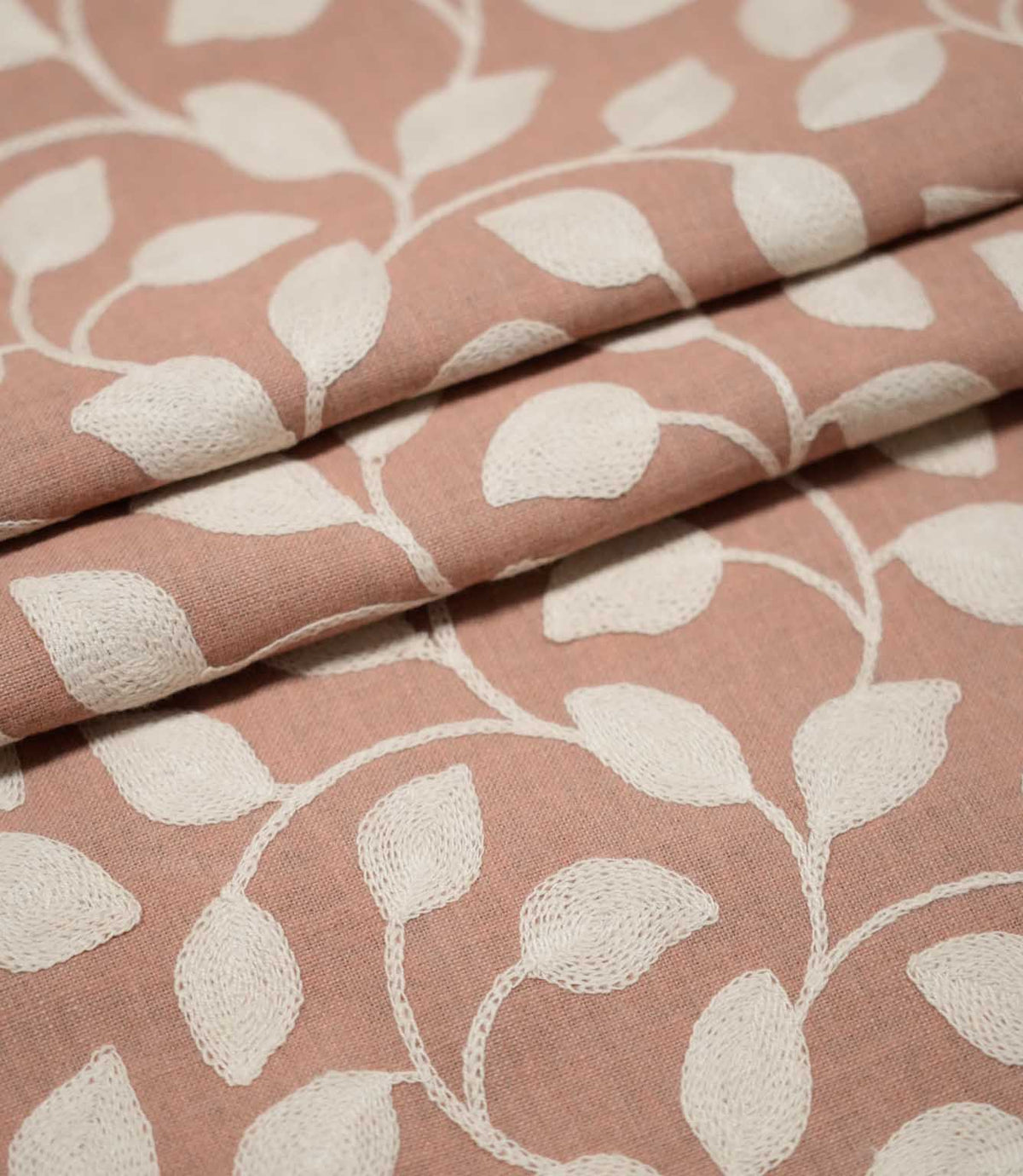 Ellerton Blush embroidered fabric arranged in overlapping folds, featuring raised cream botanical leaves connected by flowing curved stems on a blush pink woven background, highlighting the repeating foliage pattern, detailed stitching and natural movement of the fabric.