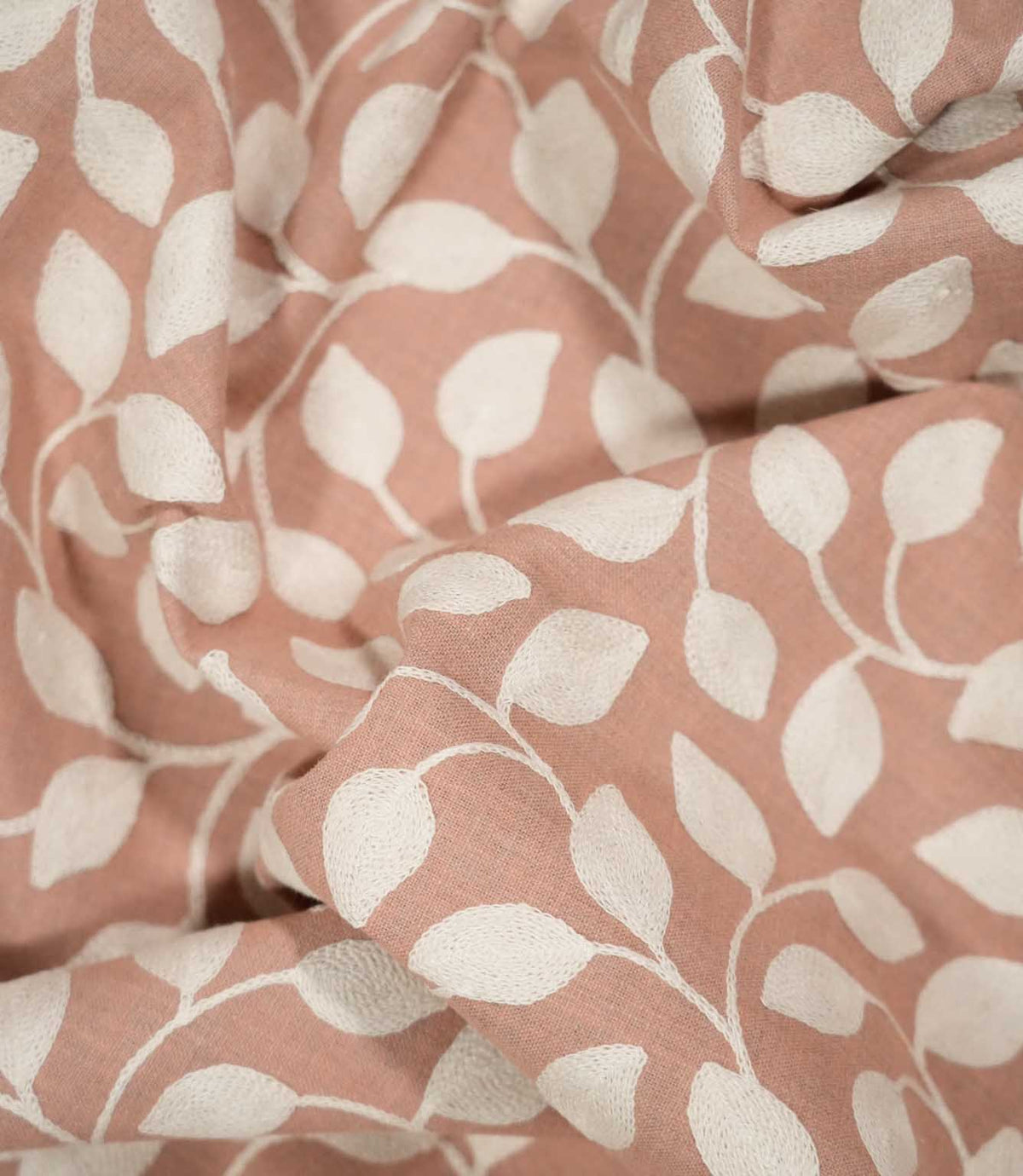Ellerton Blush fabric loosely draped into soft, flowing folds, showing the cream embroidered leaf and vine design across a warm blush pink background and demonstrating how the botanical pattern follows the fabric's natural drape and movement.
