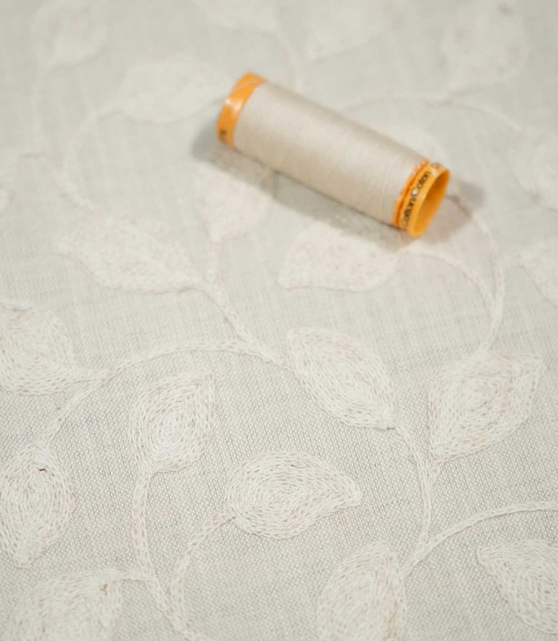 Ellerton Cloud embroidered fabric displayed with a coordinating neutral thread spool, featuring softly raised ivory leaves and flowing botanical stems across a pale cloud-grey woven background, with shallow depth of field highlighting the fabric's embroidery and textured surface.
