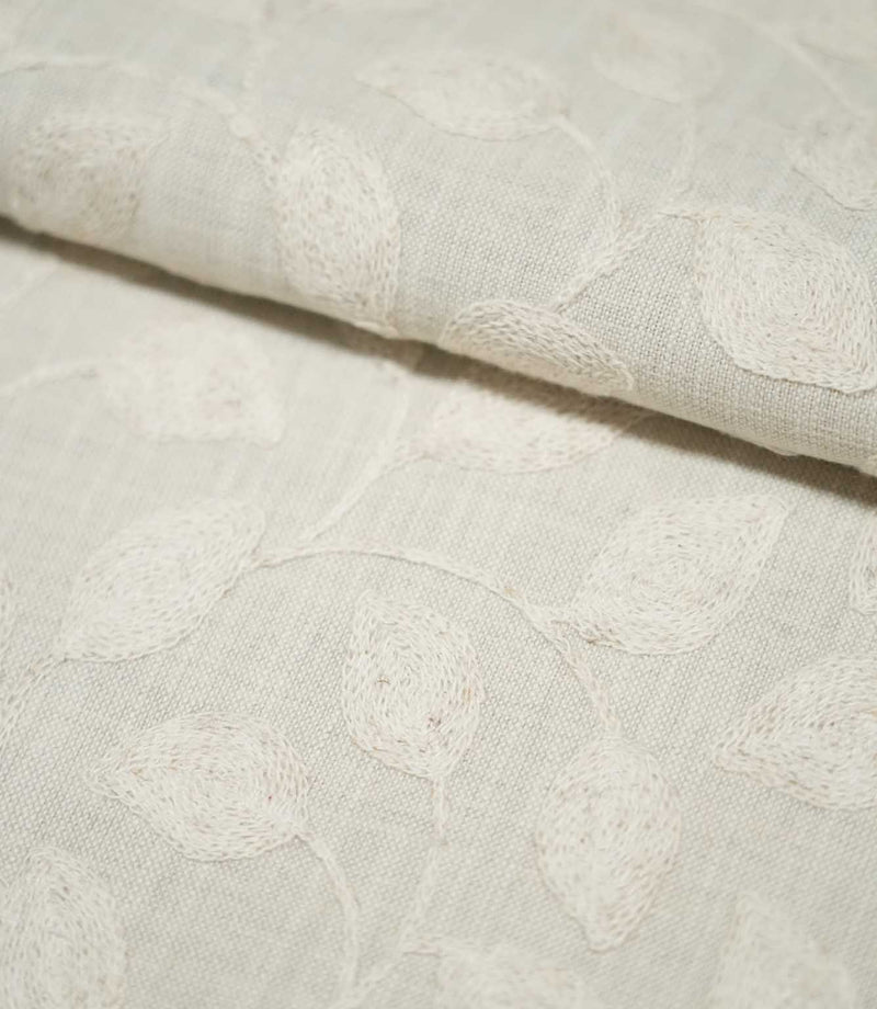 Close-up of folded Ellerton Cloud embroidered fabric showing ivory botanical leaves and gently winding stems across a pale neutral cloud-grey ground, with the soft fold highlighting the fabric's thickness, woven texture and raised embroidery.
