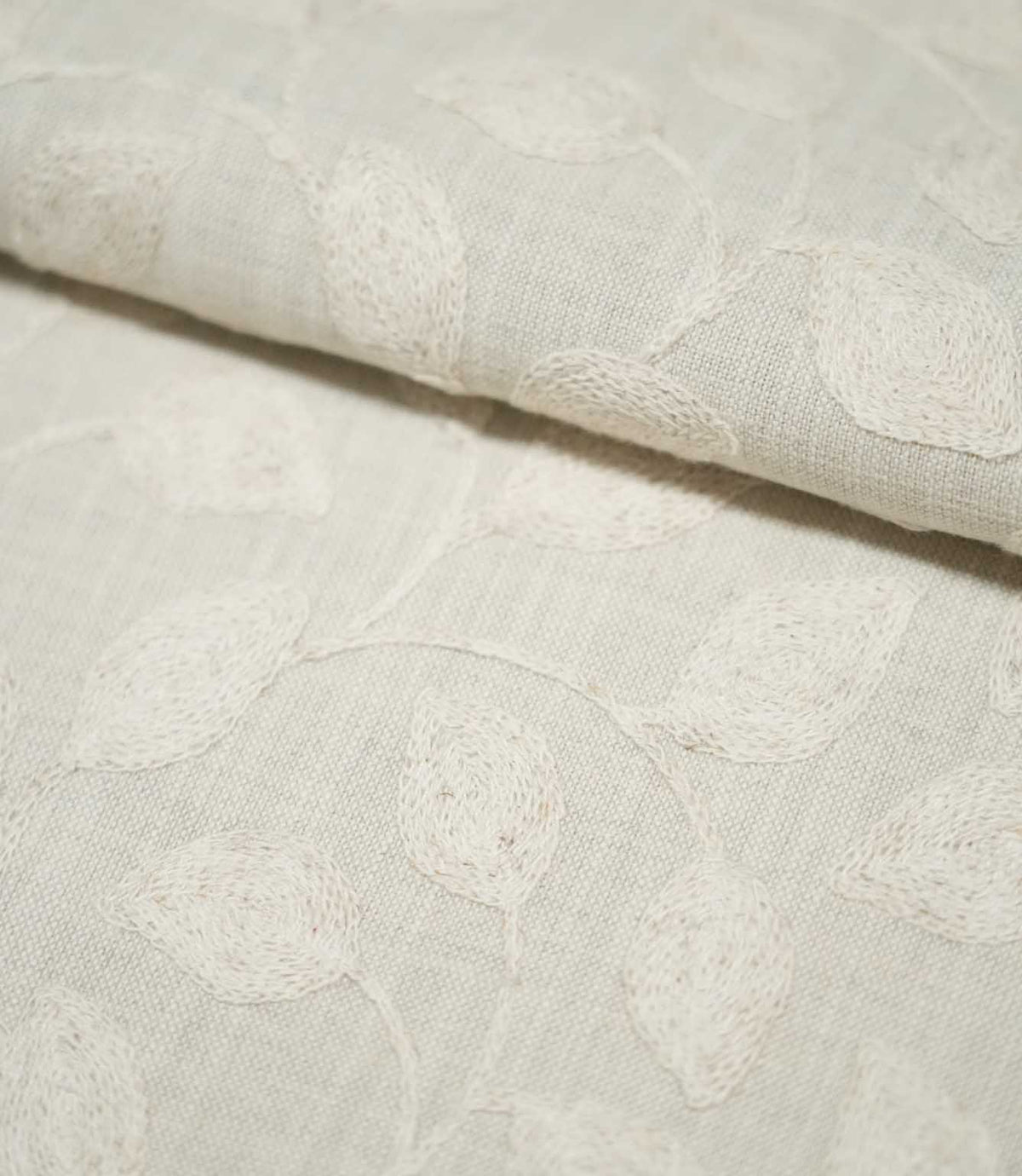 Close-up of folded Ellerton Cloud embroidered fabric showing ivory botanical leaves and gently winding stems across a pale neutral cloud-grey ground, with the soft fold highlighting the fabric's thickness, woven texture and raised embroidery.