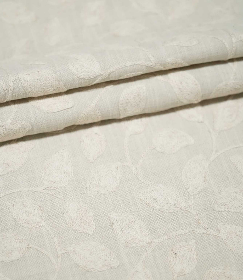 Ellerton Cloud fabric arranged in neat overlapping folds, showing the tonal ivory embroidered leaf and trailing vine pattern across a pale cloud-grey woven background, highlighting the subtle botanical design, tactile stitching and softly structured drape.