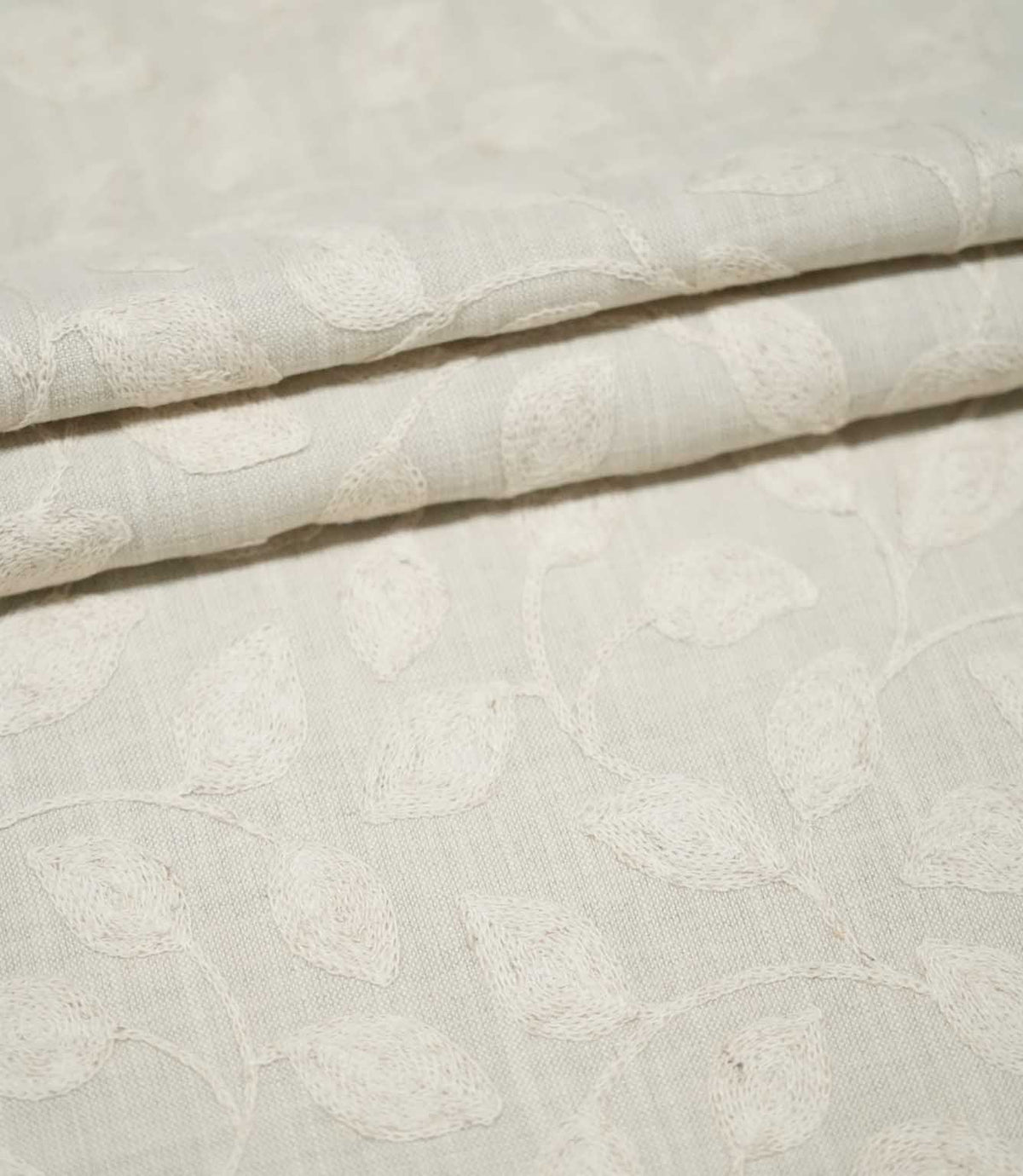 Ellerton Cloud fabric arranged in neat overlapping folds, showing the tonal ivory embroidered leaf and trailing vine pattern across a pale cloud-grey woven background, highlighting the subtle botanical design, tactile stitching and softly structured drape.