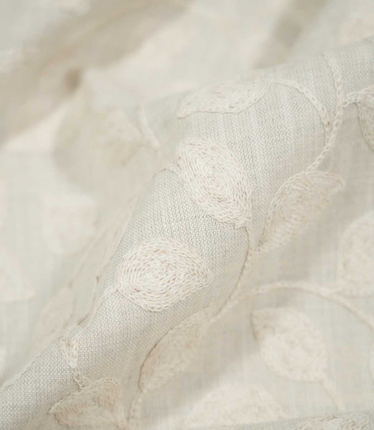 Macro close-up of softly folded Ellerton Cloud fabric, focusing on raised ivory embroidered leaves and delicate curving stems against the pale cloud-grey woven background, with shallow depth of field emphasising the detailed stitching, weave and tactile texture.